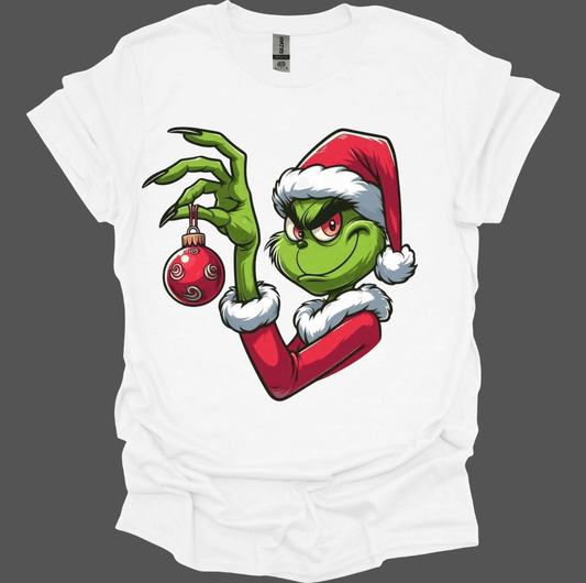White Christmas With the Grinch Tee with a graphic of a green character holding a red ornament, wearing a Santa hat.